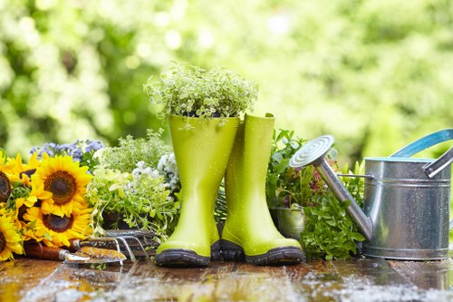 The image displays a garden scene with a dark wooden outdoor table set against a background of lush, green foliage. On the table, there are vibrant sunflower heads and various potted plants, adding bright yellow and green colours to the scene. Prominently placed in the centre are a pair of bright green rain boots filled with small white flowers, serving as unconventional plant containers. To the right of the boots, there is a traditional metal watering can with a blue handle, and a garden trowel with a wooden handle rests nearby. The surface of the table appears slightly wet, indicating recent watering or rainfall, and the overall scene suggests a well-maintained garden area, typical of residential outdoor spaces in Hatch End, where gardening and lawn care services are commonly provided by companies such as Gardening Services Hatch End. The lighting is natural, with soft sunlight filtering through the greenery, creating a fresh and inviting atmosphere for outdoor gardening activities.