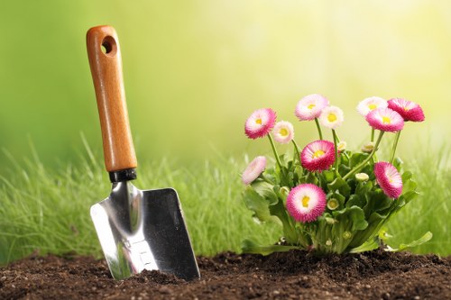 A close-up view of a garden bed in a residential outdoor space, featuring a freshly dug patch of dark, fertile soil with a small, metal hand trowel that has a wooden handle inserted into the ground, suggesting recent planting activity. To the right, there is a vibrant cluster of pink and white daisy-like flowers with round petals and yellow centers, growing amidst green foliage. The background displays a softly blurred grassy lawn, indicating a well-maintained garden area, with natural daylight illuminating the scene and highlighting the textures of the soil, plants, and gardening tools. This image reflects typical garden maintenance and planting activities that a professional gardening service such as Gardening Services Hatch End might undertake, situated within a local residential setting around HA5 postal code, in Hatch End, enabling a natural connection to local outdoor care and landscaping services.