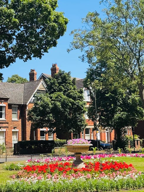 A peaceful residential street scene featuring a well-maintained garden area in the foreground with lush, green grass and a paved pathway. Two black waste bins are situated on the grass near the path, 