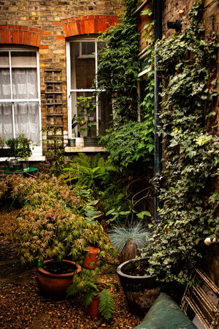 A small, charming brick building with a dark grey tiled hipped roof and two brick chimneys, situated behind a black wrought iron fence. The house has two white-framed sash windows on either side of a